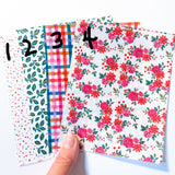 A hand holds a fan of patterned paper sheets on a white background. The papers show different designs: a white sheet with green leaves, a white sheet with blue and pink plaid, a white sheet with red and orange plaid, and a white sheet with pink and red flowers and green leaves.